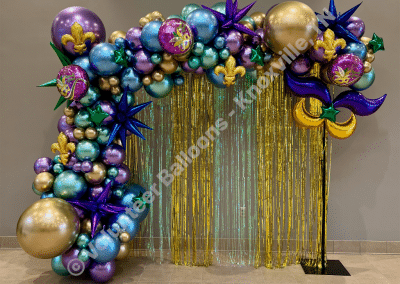 Mardi Gras themed balloon arch with a foil fringe backdrop, colors are purple, green, gold, blue