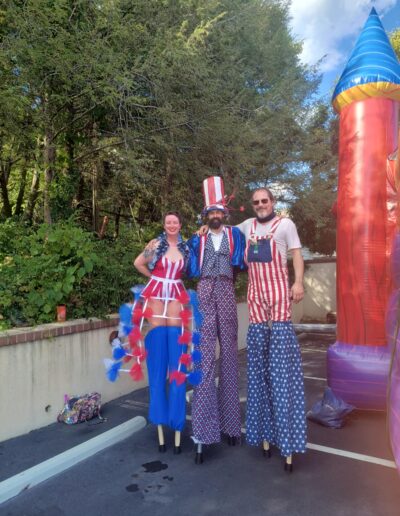 Three performers stand on stilts in fourth of July themed outfits in front of a bouncy castle